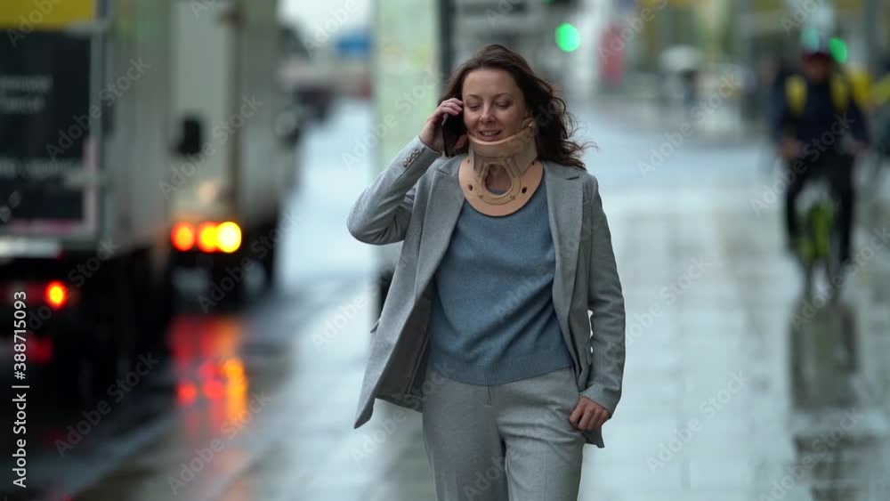 A middle-aged woman in a jacket walks down a street in the city on an autumn day against the background of a highway and talks on the phone, she has a corset around her neck. A middle-aged woman in a jacket walks down a street in the city on an autumn day against the background of a highway and talks on the phone, she has a corset around her neck.