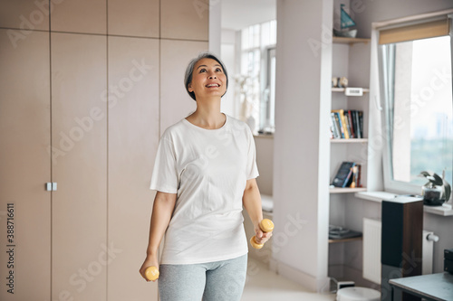 Adult Asian female in white shirt holding sport tools in hands while looking up in living room