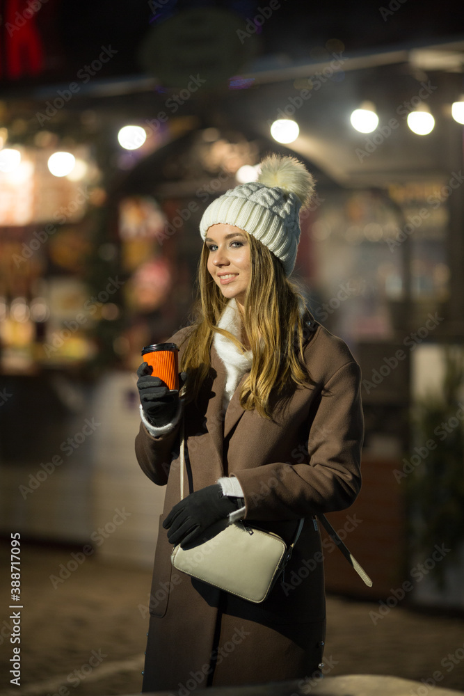 a blonde girl, in the evening, against the background of the lights of the fair with a drink in an orange glass, a white hat and a beige coat walks on the street.