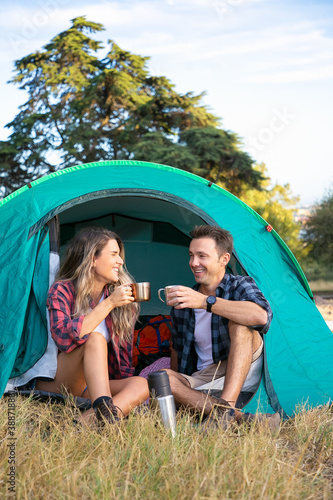 Cheerful couple sitting in tent, chatting and drinking tea. Happy hikers relaxing on lawn, camping and enjoying nature. Travelers outdoors in nature. Tourism, adventure and summer vacation concept