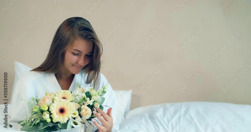 Happy lady in bathrobe holds fresh flowers bouquet sitting on large comfortable bed in hotel room in sunny morning Happy lady in bathrobe holds fresh flowers bouquet sitting on large comfortable bed in hotel room in sunny morning