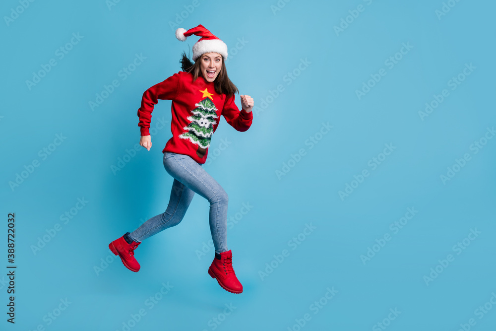 Full length profile photo of charming cute lady jump sprinting air open mouth wear santa x-mas headwear red ornamented pullover jeans boots isolated blue color background Full length profile photo of charming cute lady jump sprinting air open mouth wear santa x-mas headwear red ornamented pullover jeans boots isolated blue color background