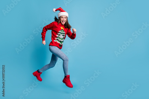 Full length profile photo of charming cute lady jump sprinting air open mouth wear santa x-mas headwear red ornamented pullover jeans boots isolated blue color background