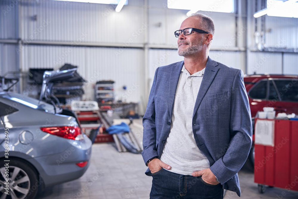 Joyful bearded man standing in vehicle repair shop Joyful bearded man standing in vehicle repair shop