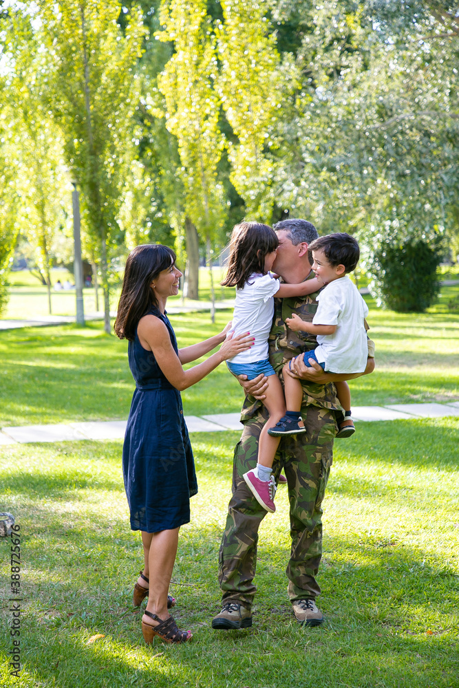 Fototapeta premium Cheerful mom and two kids meeting military dad in camouflage uniform outdoors. Father taking children in arms and kissing. Family reunion or returning home concept