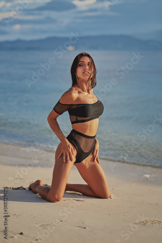 Obraz na płótnie beautiful sexy woman in black swimwear posing at the beach and looking at camera while sunset w salonie