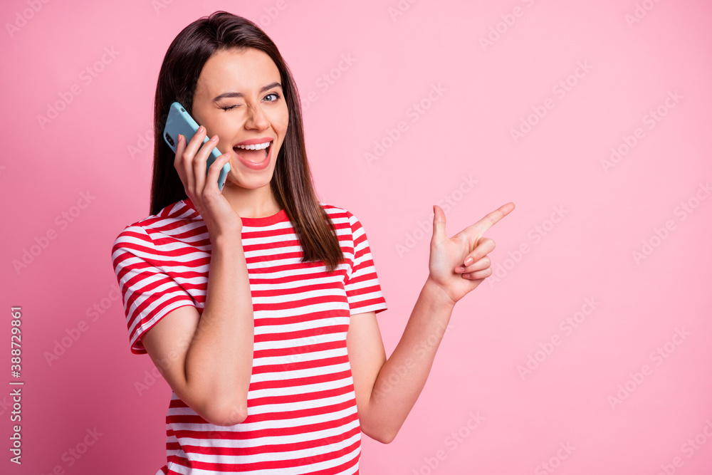 Photo portrait of girl talking on cell phone pointing finger at blank space winking isolated on pastel pink color background Photo portrait of girl talking on cell phone pointing finger at blank space winking isolated on pastel pink color background