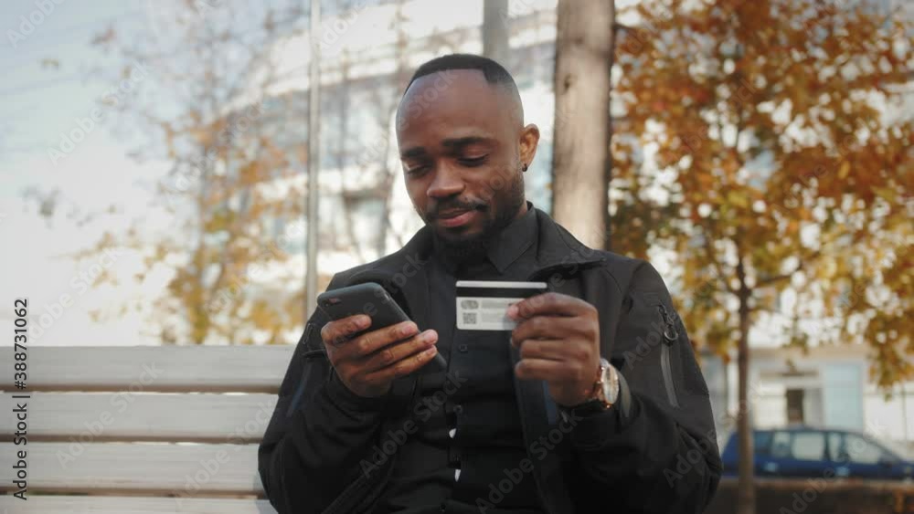 Smiling customer holding credit card and smartphone sitting on bench in the park. Happy african american man shopper using instant easy mobile payments making purchase in online store.