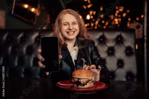 Portrait of a young lovely hungry woman sitting in the street fast junk food restaurant cafe hold showing smartphone mock up screen and eating enjoying a fresh tasty burger on a red plate. 