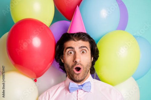 Portrait of shocked man open mouth wear cone cap pink shirt purple bow isolated many balloons creative background