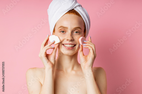 Pretty glad young caucasian woman with towel on head, bare shoulders, holding two cosmetic sponges on cheeks, enjoying beauty day smiling broadly posing against pink studio wall. Skin care.