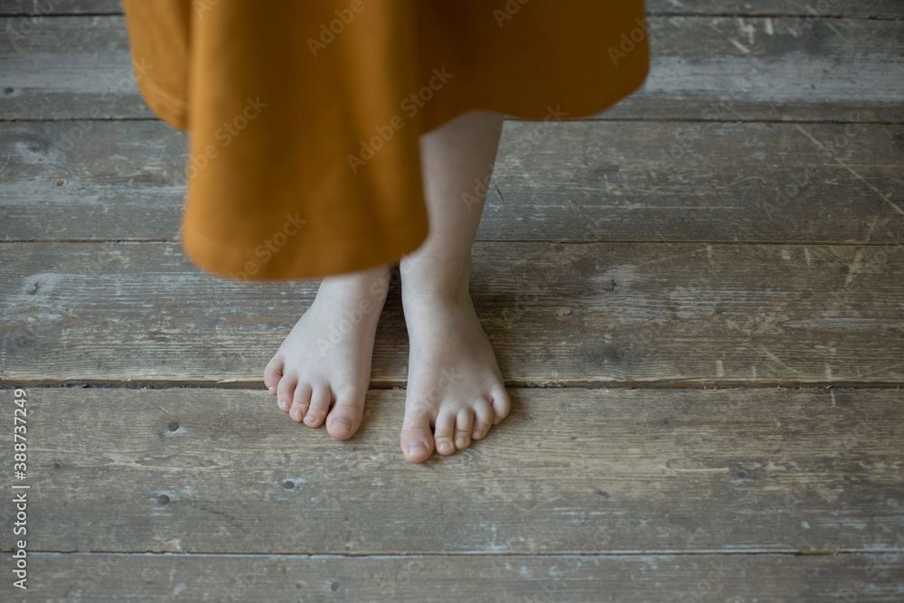 Child's feet are on the wooden floor. Little girl in orange linen dress standing on the floor by bare foot. Childhood. Image with selective focus. Wooden background. Child's feet are on the wooden floor. Little girl in orange linen dress standing on the floor by bare foot. Childhood. Image with selective focus. Wooden background.