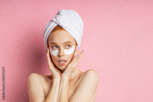 Attractive nude young African woman applying under eye patches to reduce dark circles and puffiness, keeping palms on cheeks, standing against pink background with copy space for text.
