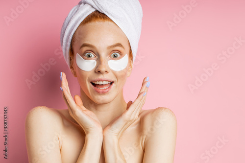 Closeup of surprised cheerful redhead young woman applying moisturizing hyaluronic patches, biting lips, looking to side with happy expression posing over pink background with copy space.