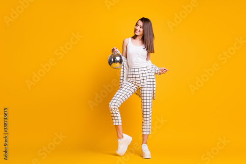 Full size photo of cheerful nice brown hair girl hold disco ball look wear suit tank-top sneakers isolated over yellow background