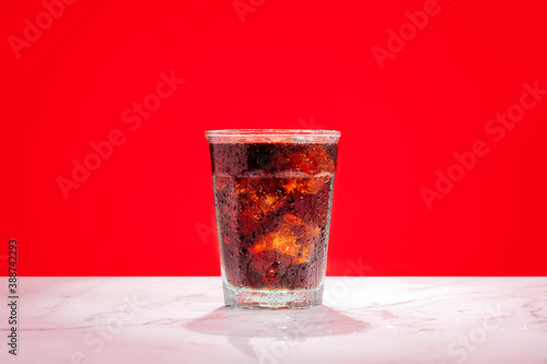 Ice cold glass of soda, cola, fizzy drink, carbonated beverage sits against a red wall on a marble surface with ice cubes and condensation on the glass making it refreshing and inviting.
