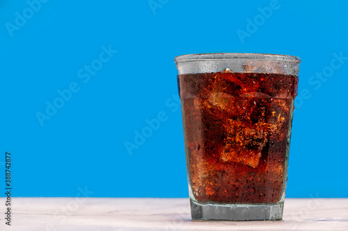 Ice cold glass of soda, cola, fizzy drink, carbonated beverage sits against a blue wall with ice cubes and condensation on the glass making it refreshing and inviting.