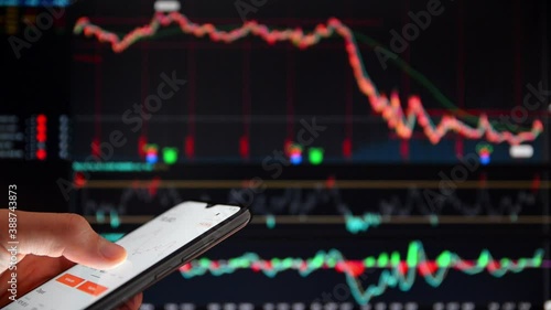 Investor analyzing the stock prices on a smartphone. Person with smartphone in front of a big screen with falling share prices. Dropping stock prices on a screen. Falling market rates on a screen.