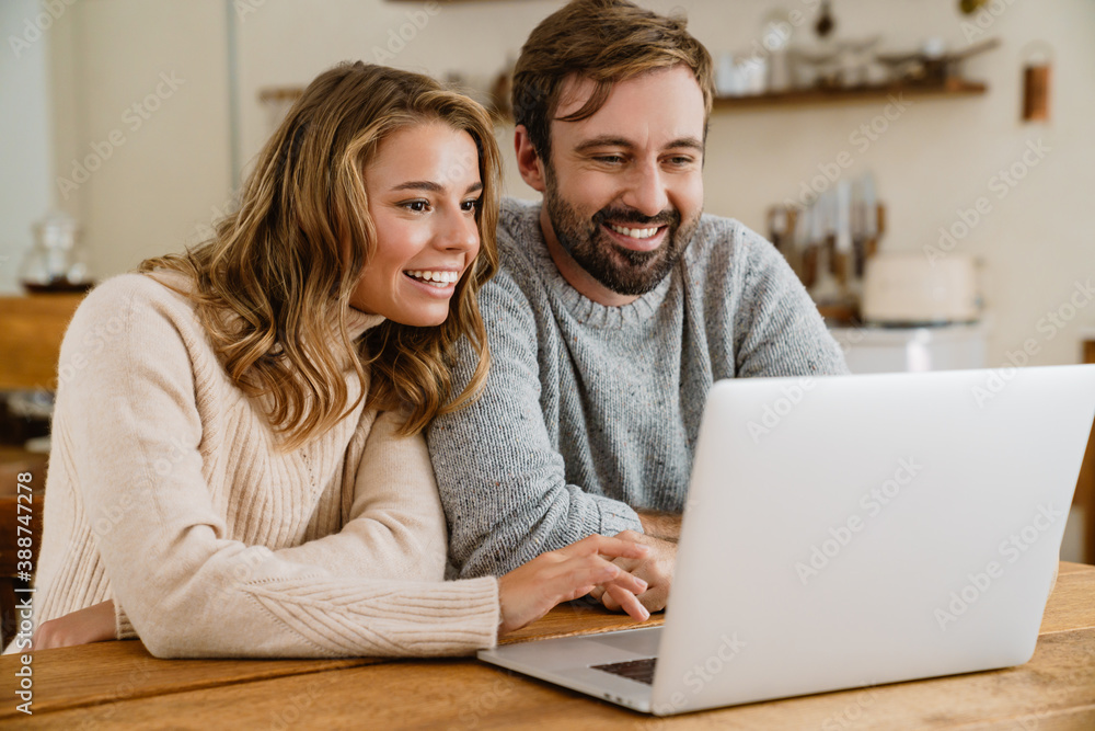 Beautiful happy couple using laptop while sitting in kitchen Beautiful happy couple using laptop while sitting in kitchen