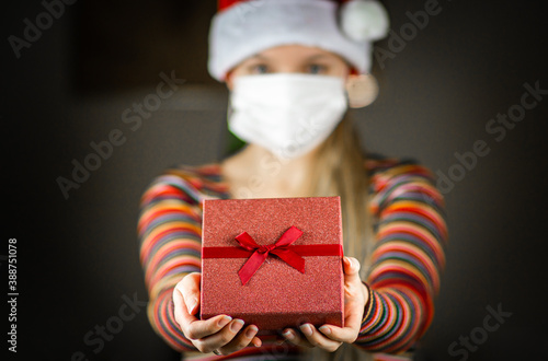 Young blonde caucasian girl in Santa hat and striped jumper , with protective mask on face, holding, giving a gift, present to the camera.  Coronavirus lockdown Christmas concept.