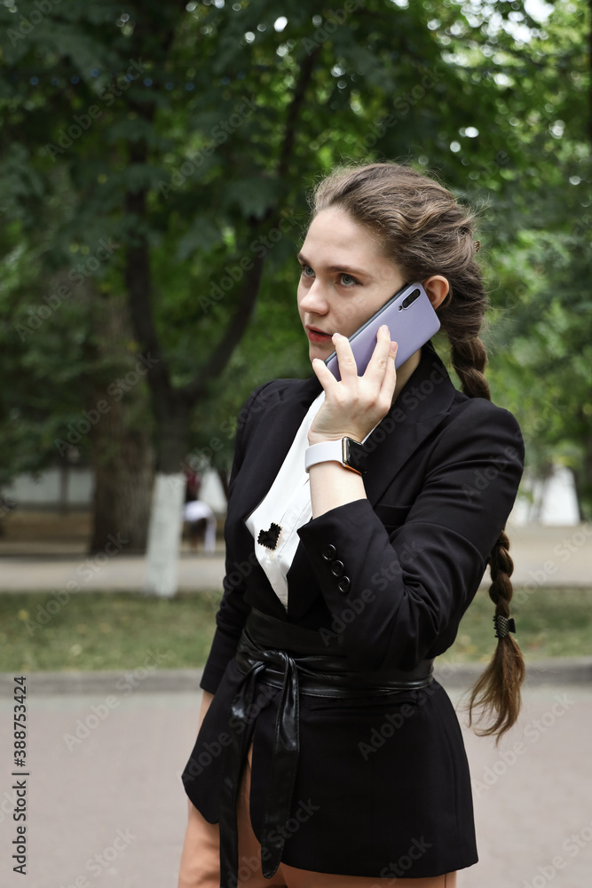 Young businesswoman in suit walks along the alley next to the road and seriously talks on smartphone on summer day Young businesswoman in suit walks along the alley next to the road and seriously talks on smartphone on summer day