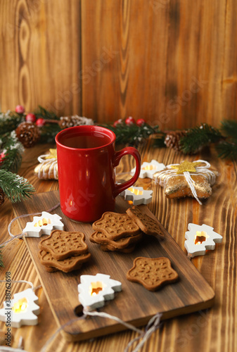 A red Cup of fragrant tea and homemade ginger cookies stand on a wooden Board among the branches of a fir tree with cones and red berries and lights in the shape of Christmas trees.Selective focus