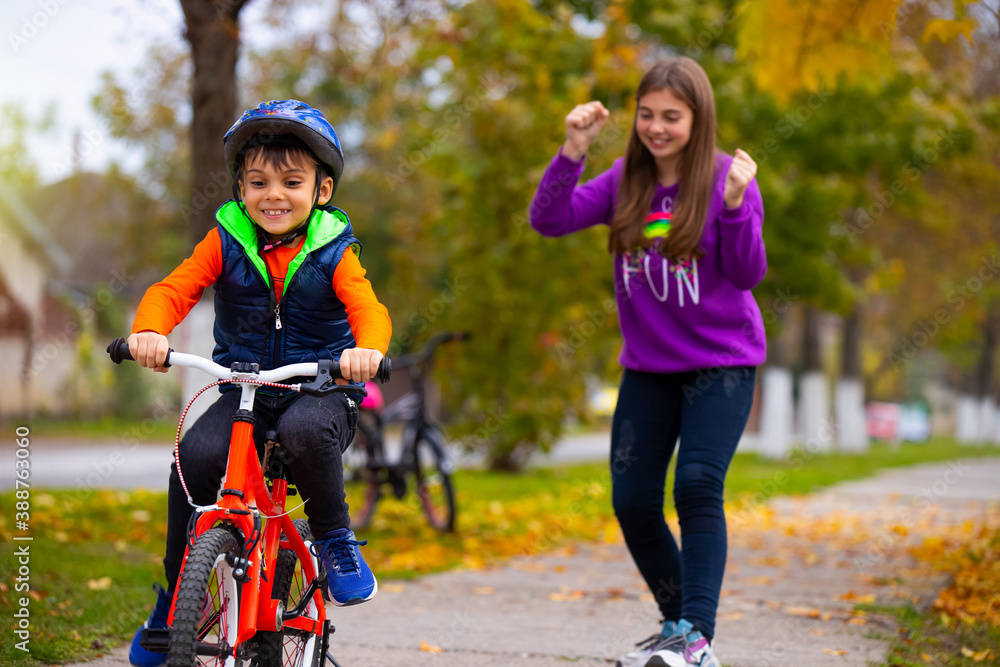 Brother and sister in the park. The boy learns to ride a bike alone. The sister in the background rejoices over his success. Photo on the background of autumn. Brother and sister in the park. The boy learns to ride a bike alone. The sister in the background rejoices over his success. Photo on the background of autumn.