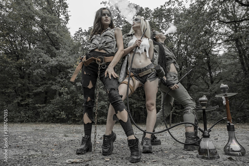 Three girls in torn trash clothes in an apocalyptic manner smoke a hookah. © baxys