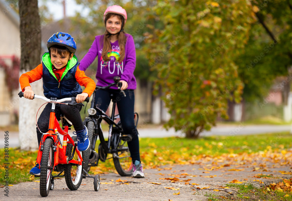 Fototapeta premium Brother and sister in the park. Boy rides a bike. A girl in the background with a bicycles on a background of autumn. Photo with side space