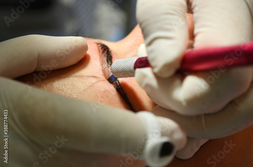 Woman lying on procedure of microblading. Cosmetologist in white gloves makes permanent makeup on the eyebrows.