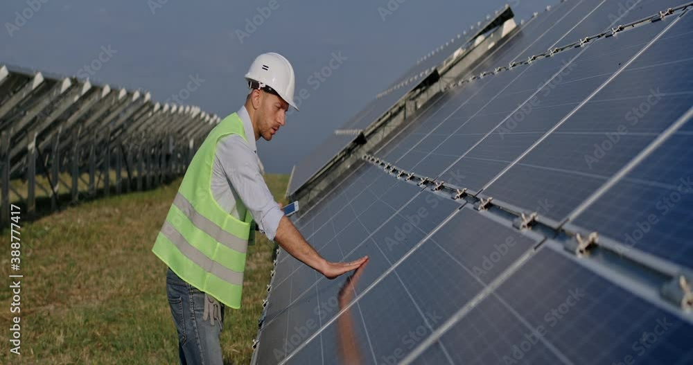 Male engineer entering data while using tablet and touching solar panel. Man in uniform checking work of photovoltaic cell and smiling while standing green energy farm. Anamorphic lens. Male engineer entering data while using tablet and touching solar panel. Man in uniform checking work of photovoltaic cell and smiling while standing green energy farm. Anamorphic lens.