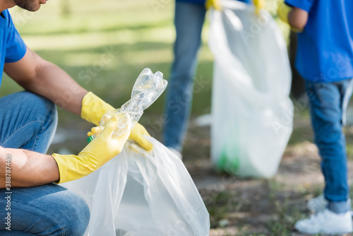 cropped view of man holding plastic bottle and recycled bag near family on blurred background, ecology concept photo