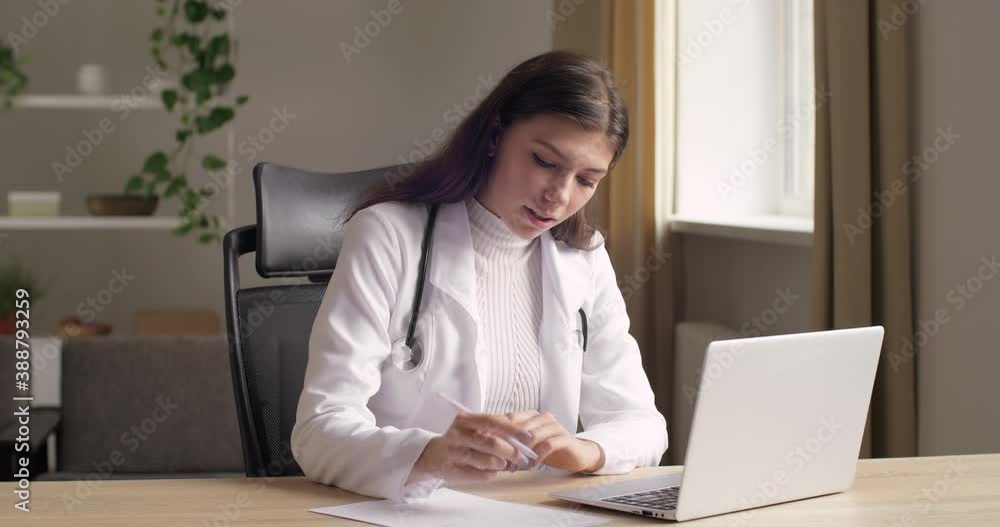 Young smart friendly female clinician nurse in white medical gown using laptop for remote patient consultation in hospital office in epidemic of covid. Online medicine, distant remote doctor concept Young smart friendly female clinician nurse in white medical gown using laptop for remote patient consultation in hospital office in epidemic of covid. Online medicine, distant remote doctor concept