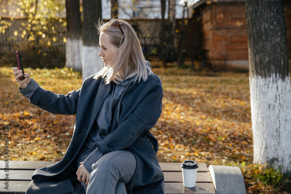 Beautiful young happy 30-year-old woman takes a selfie with her smartphone on a bench in an autumn Park. The concept of the season, technology and people . Beautiful young happy 30-year-old woman takes a selfie with her smartphone on a bench in an autumn Park. The concept of the season, technology and people .