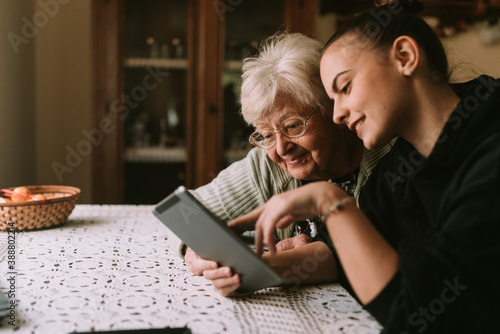 Smiling caucasian grandmother and beautiful granddaughter in the kitchen looking at the tablet. The granddaughter teaches her grandmother to use new technology