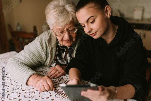 The senior caucasian grandmother and her beautiful teenage granddaughter look at the pictures on the tablet in the room