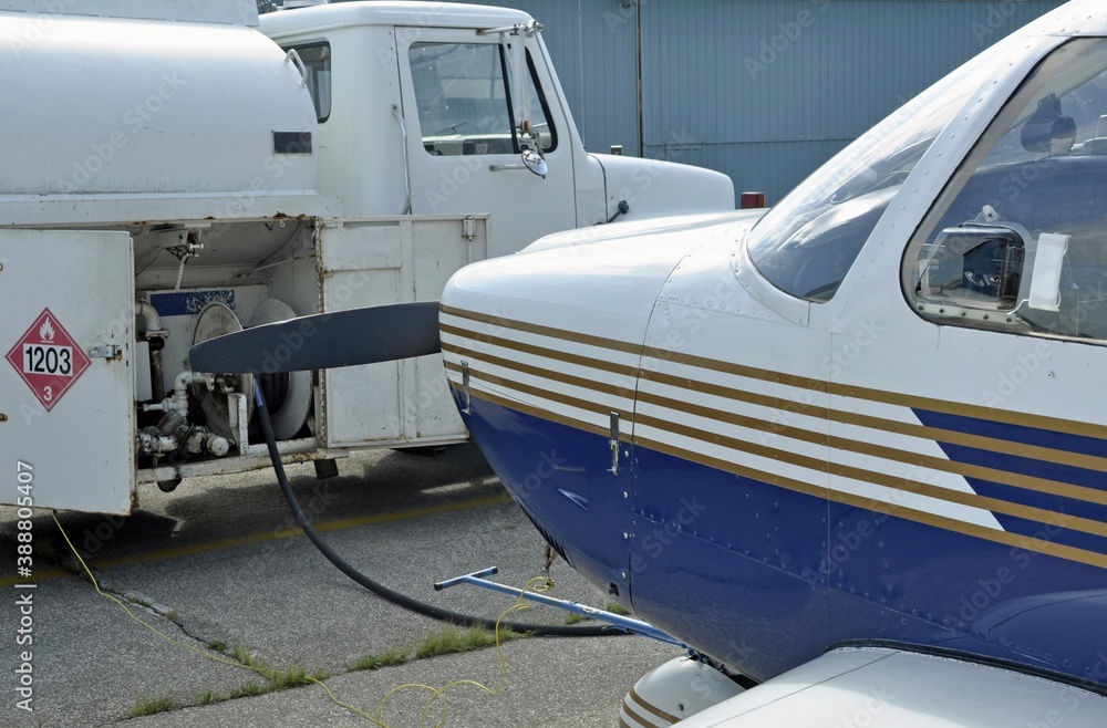 Closeup of a blue and white airplane being refueled by a nearby white fuel truck with an 1203 gasoline or petrol flammable liquid placard