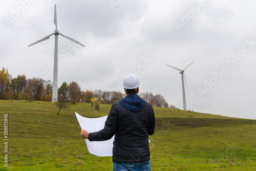Back view of male engineer wears white helmet holding plan at wind turbine autumn cloudy sky meadow