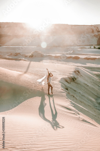 young woman in white body is standing in gymnastic pose on the sand in desert at sunset, fashion sport concept, free space 