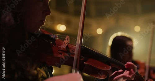 Cinematic close-up shot of female violinist is playing violins during musical concert on classic theatre stage with symphony orchestra performing on background. photo