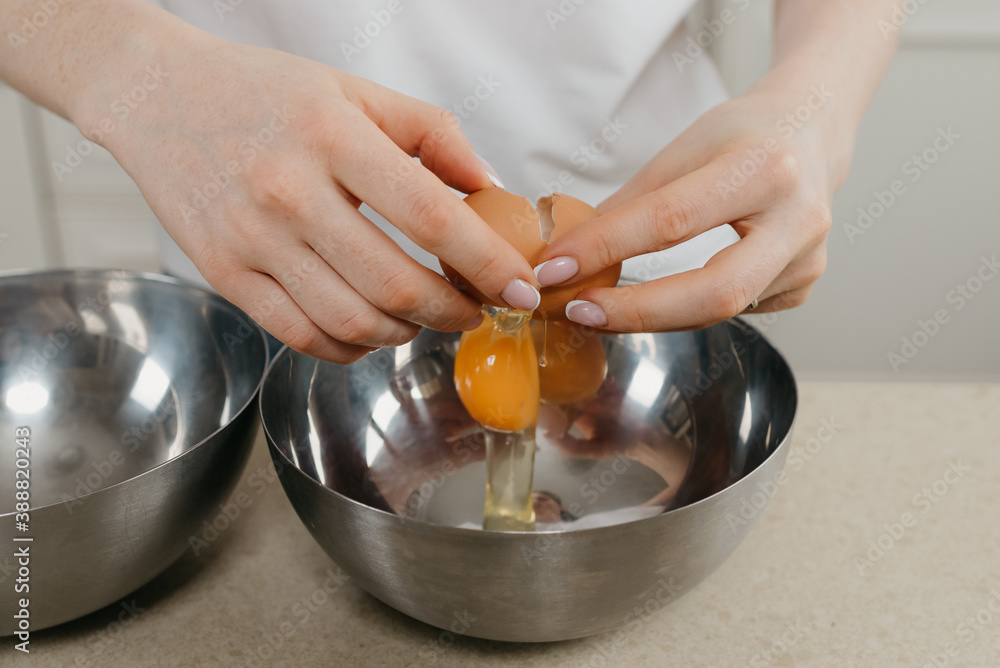 A close photo of the hands of a young woman who is breaking the organic farm egg above the stainless steel soup bowl in the kitchen. A close photo of the hands of a young woman who is breaking the organic farm egg above the stainless steel soup bowl in the kitchen.