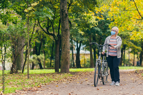senior woman walks in the park with a bicycle in a protective medical mask