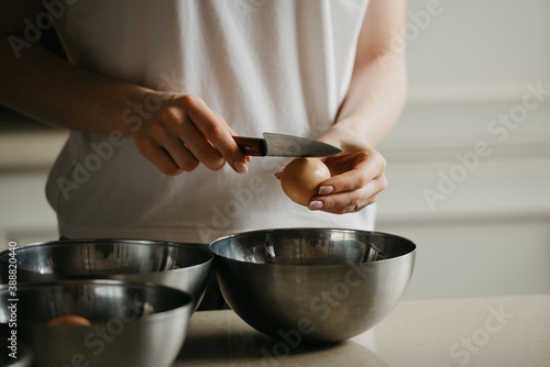 A dark photo of the hands of a young woman who is breaking the eggshell of the organic farm egg with the knife above the stainless steel soup bowl in the kitchen