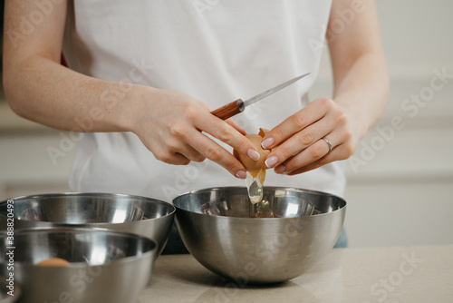 A close photo of the hands of a girl who is breaking the organic farm egg holding a knife above the stainless steel soup bowl in the kitchen.