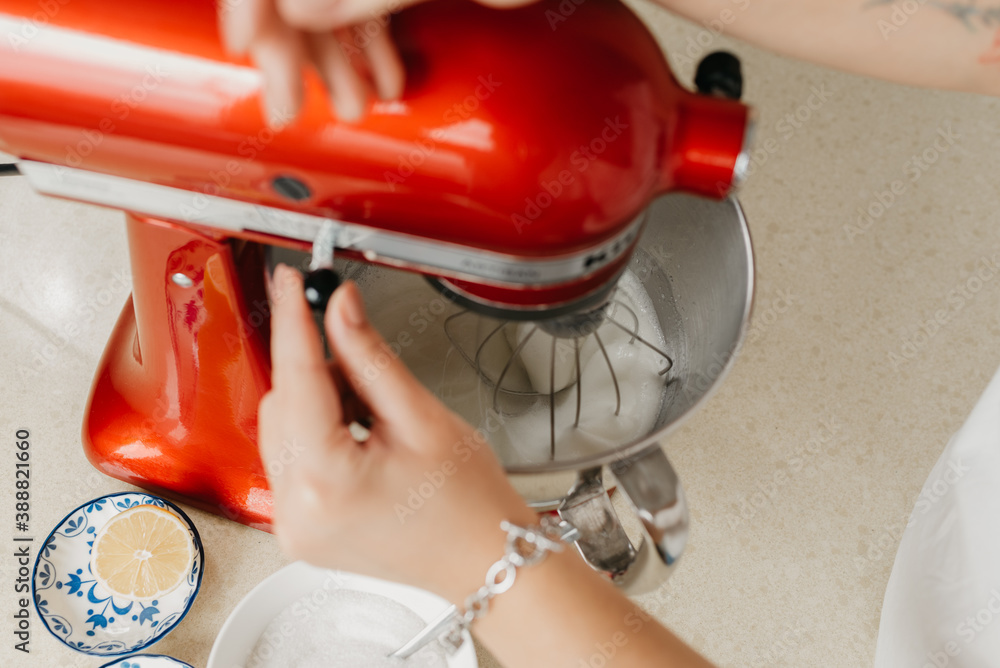 The hands of a woman who is switching speeds of a red stand mixer which is whipping the egg whites in a stainless steel soup bowl in a kitchen. The hands of a woman who is switching speeds of a red stand mixer which is whipping the egg whites in a stainless steel soup bowl in a kitchen.