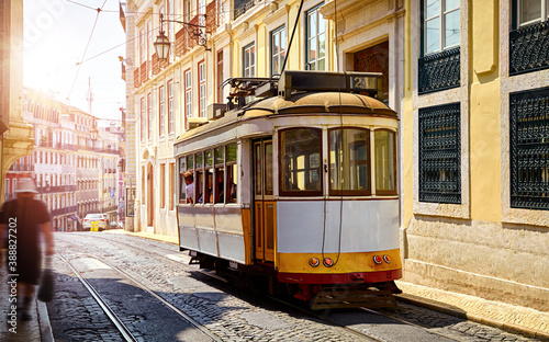 Lisbon Portugal. Yellow vintage tram driving by street of paving stones in district Alfama. Cityscape panorama with old houses and tower in sunny day with blue sky and white clouds.
