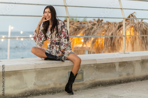 Outdoor, evening, young italian woman brunette and long hair with flowered dress sitting on the wall of a beach of a seaside city in the act of nightlife