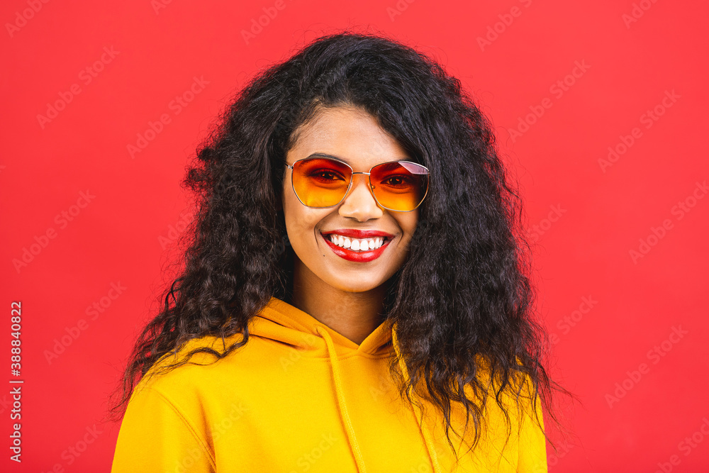 Portrait of beautiful positive african american black curly woman in sunglasses standing isolated over red background.