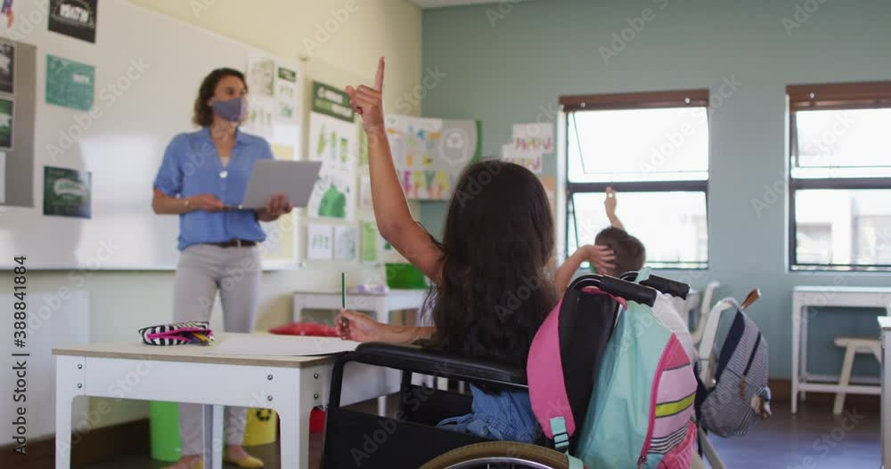 Disable girl wearing face mask raising her hand while sitting on wheelchair in class Disable girl wearing face mask raising her hand while sitting on wheelchair in class