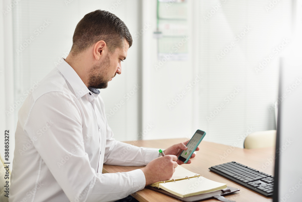 Young male office manager in the office looks carefully at the smartphone and writes in the notebook Young male office manager in the office looks carefully at the smartphone and writes in the notebook
